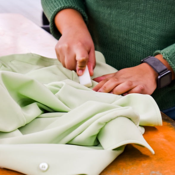 A person wearing a green sweater uses tailor’s chalk on light green fabric, showing the care often needed when using Meliora’s Laundry Stain Stick for toddler clothes stain removal before sewing.