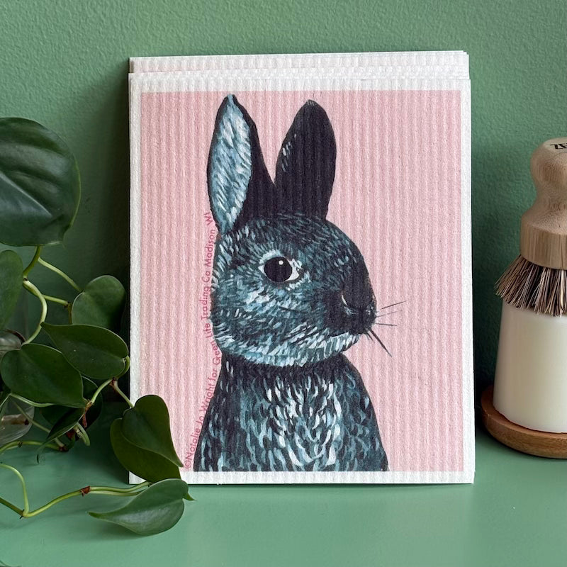 Dishcloth with a rabbit illustration on a pink background, placed on a green surface with a plant and bottle in the background.