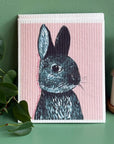 Dishcloth with a rabbit illustration on a pink background, placed on a green surface with a plant and bottle in the background.