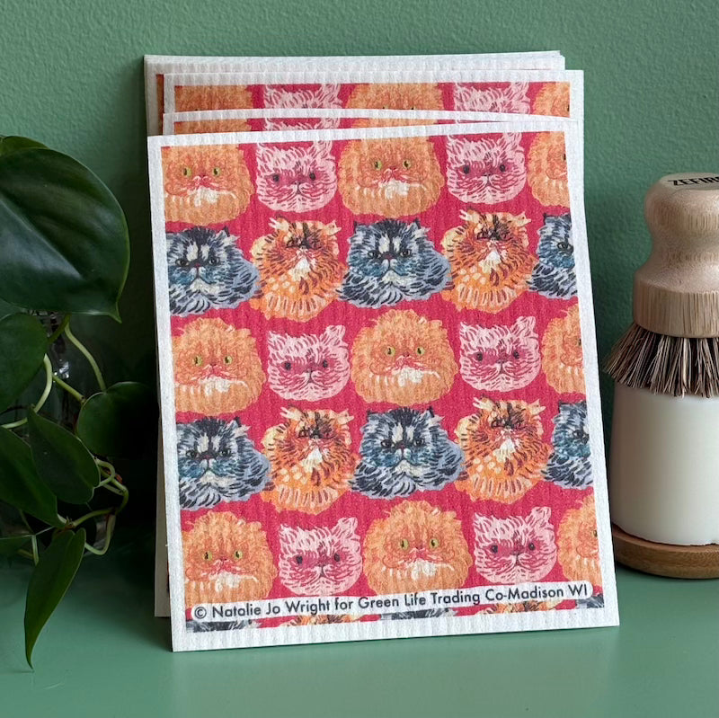 Patterned towel with cat faces on a green surface with a plant and bottle in the background
