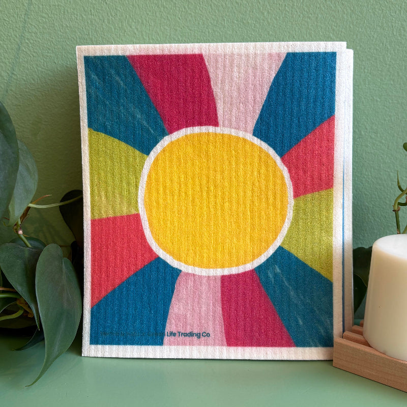 A colorful Swedish dishcloth with a sun design on it by local Madison Artist Bernie & Zuzu. It is sitting on a green background by a plant and solid dish puck.