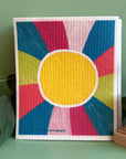 A colorful Swedish dishcloth with a sun design on it by local Madison Artist Bernie & Zuzu. It is sitting on a green background by a plant and solid dish puck.