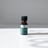 A small amber glass bottle of Mountain Rose Herbs Tea Tree Essential Oil (certified organic, Melaleuca alternifolia) with a black cap sits on a white surface, casting a shadow in bright natural light.