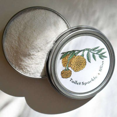 A round metal tin of white powder is displayed with a partially open lid showing yellow flowers, green leaves, and the text Toilet Sparkle by Shade Tree Naturals.