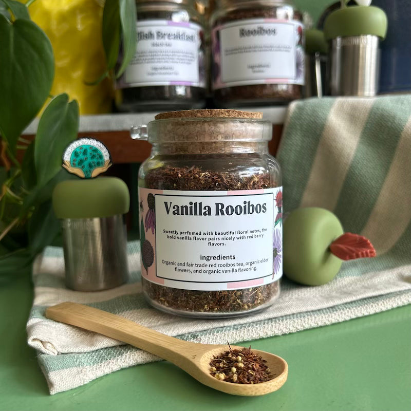A refillable jar of Vanilla Rooibos tea. The jar has a cork lid and an attachable bamboo spoon. The spoon is in front of the jar with vanilla rooibos tea in it. 