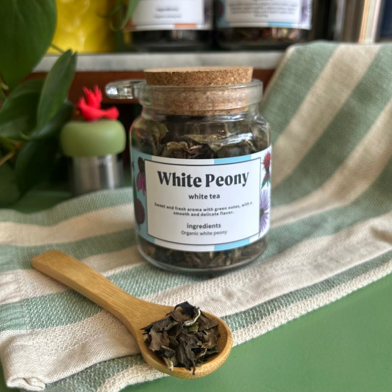 A refillable jar of white peony tea. The jar has a cork lid and an attachable bamboo spoon. The spoon is in front full of tea. 