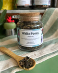 A refillable jar of white peony tea. The jar has a cork lid and an attachable bamboo spoon. The spoon is in front full of tea. 