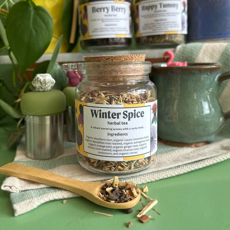 A refillable jar of winter spice tea. The jar has a cork lid with an attachable bamboo spoon. The spoon is in front of the jar with winter spice tea in it. 