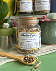 A refillable jar of winter spice tea. The jar has a cork lid with an attachable bamboo spoon. The spoon is in front of the jar with winter spice tea in it. 