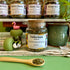 A refillable glass jar of Yerba Mate herbal tea with a cork lid sits on a striped cloth on a green surface, with a wooden spoon of loose tea in front and other tea jars  in the background.