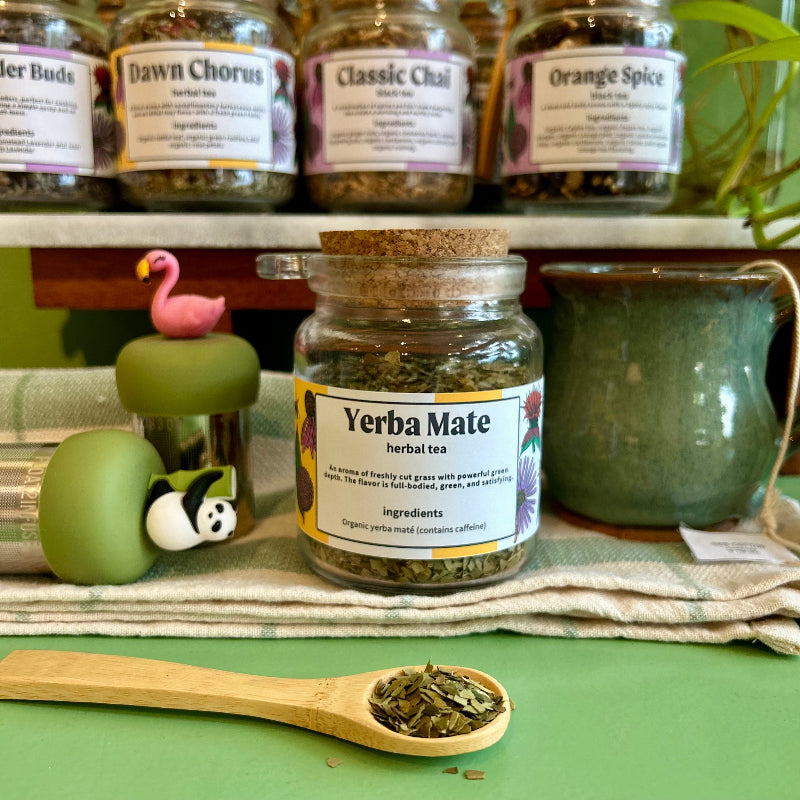 A refillable glass jar of Yerba Mate herbal tea with a cork lid sits on a striped cloth on a green surface, with a wooden spoon of loose tea in front and other tea jars  in the background.