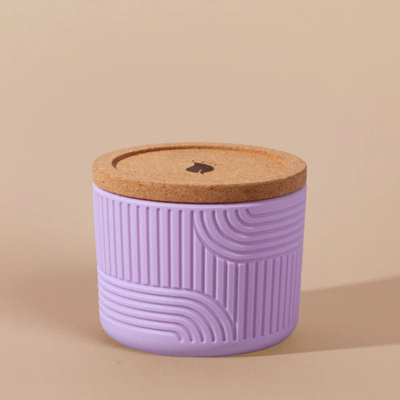 The Arbor Made Refillable Candle Jar in the silicone jar style and in the color lilac. The jar features a cork lid with a small leaf design on it and a silicone body with a linear geometric pattern on it. The Jar is siting on a beige background.