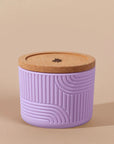 The Arbor Made Refillable Candle Jar in the silicone jar style and in the color lilac. The jar features a cork lid with a small leaf design on it and a silicone body with a linear geometric pattern on it. The Jar is siting on a beige background.