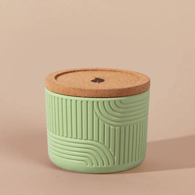 The Arbor Made Refillable Candle Jar in the silicone jar style and in the color meadow. The jar features a cork lid with a small leaf design on it and a silicone body with a linear geometric pattern on it. The Jar is siting on a beige background. 