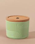 The Arbor Made Refillable Candle Jar in the silicone jar style and in the color meadow. The jar features a cork lid with a small leaf design on it and a silicone body with a linear geometric pattern on it. The Jar is siting on a beige background. 