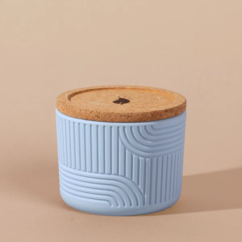 The Arbor Made Refillable Candle Jar in the silicone jar style and in the color sky. The jar features a cork lid with a small leaf design on it and a silicone body with a linear geometric pattern on it. The Jar is siting on a beige background. 