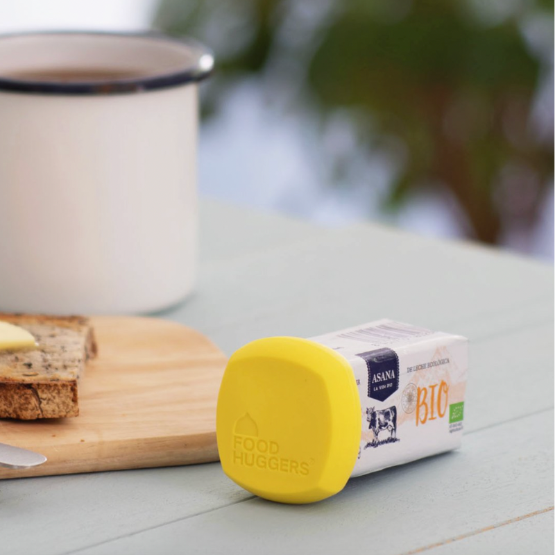 A small carton of ASANA Organic Bio Butter lies on its side on a light blue wooden table, with a bright yellow lid or cap covering the front. A white enamel mug with a black rim, toast with butter, and a knife are blurred in the background.