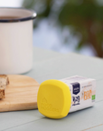 A small carton of ASANA Organic Bio Butter lies on its side on a light blue wooden table, with a bright yellow lid or cap covering the front. A white enamel mug with a black rim, toast with butter, and a knife are blurred in the background.