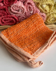 An orange hand-dyed hemp exfoliating washcloth with a basket full of rolled hemp exfoliating washcloths in the background. 