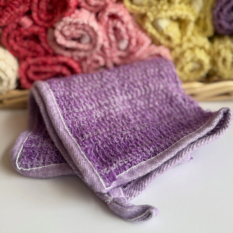 A purple hand-dyed hemp exfoliating washcloth with a basket full of rolled hemp exfoliating washcloths in the background. 
