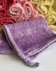 A purple hand-dyed hemp exfoliating washcloth with a basket full of rolled hemp exfoliating washcloths in the background. 