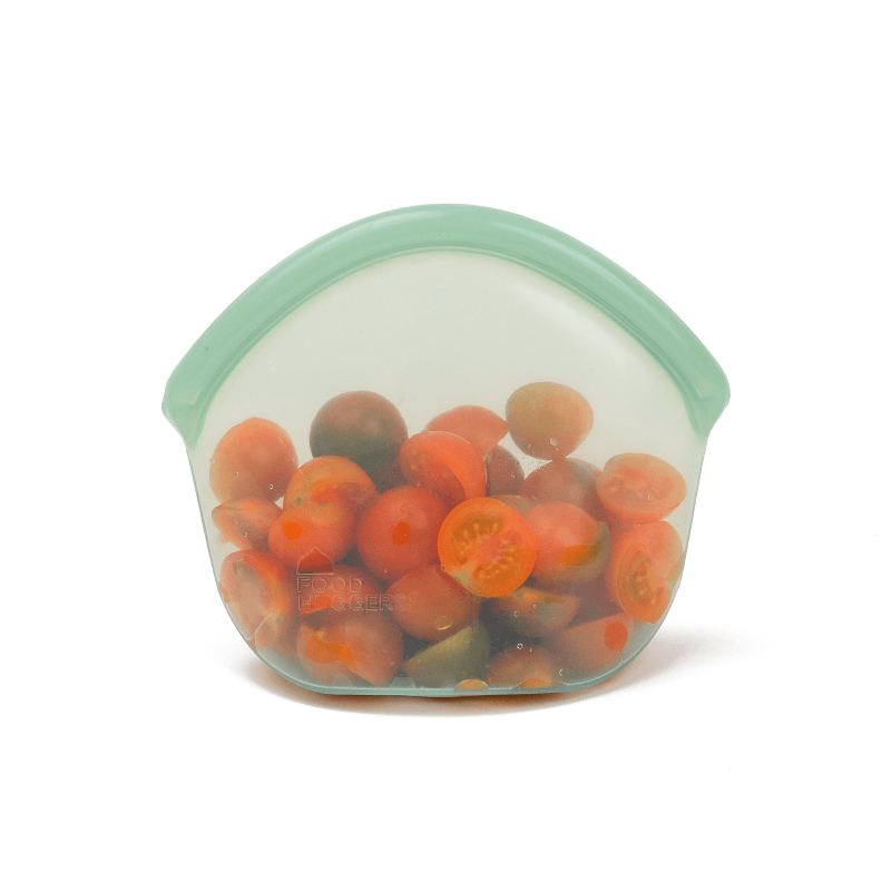 Small Green Transparent silicone reusable snack bag holding sliced tomatoes on a white background. 