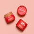 Three red Axiology Mushroom Foundation containers sit on a peach background; two jars are open to display the product, while one lid face-up completes the arrangement.