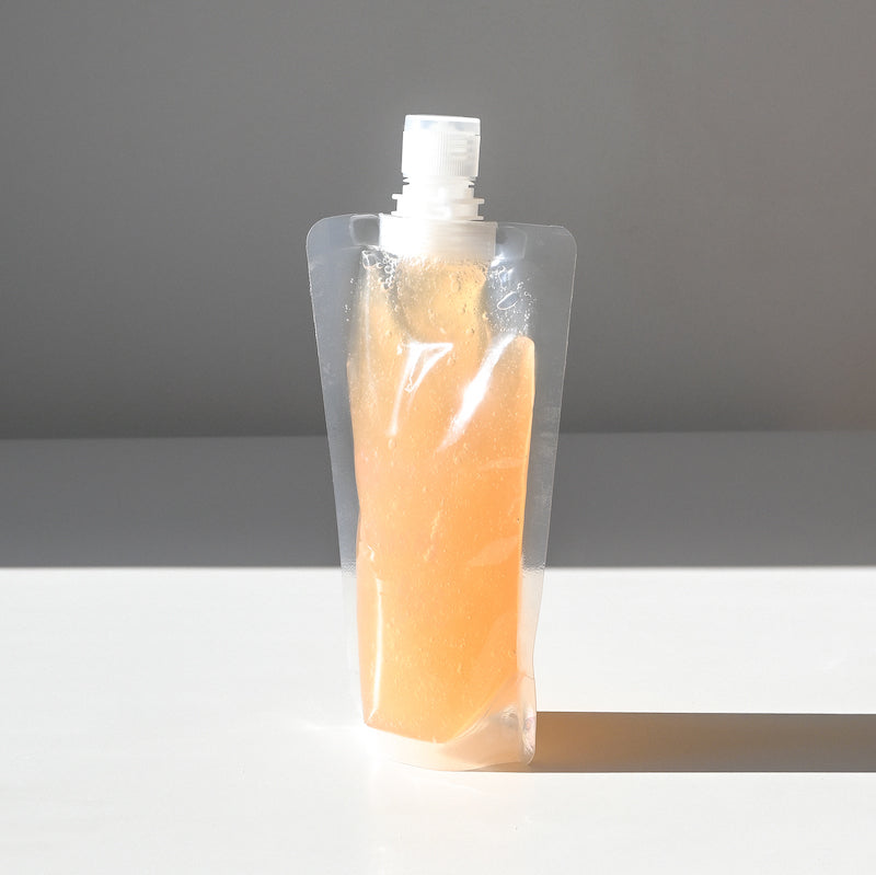 A clear refillable pouch holding orange gel face wash.