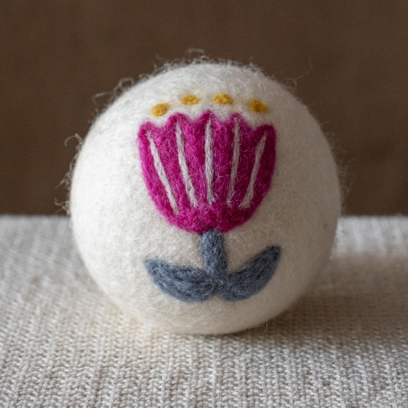 A white felted wool dryer ball featuring a purple folk art flower with a blue stem, resting on a textured beige fabric surface against a brown background.