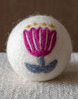 A white felted wool dryer ball featuring a purple folk art flower with a blue stem, resting on a textured beige fabric surface against a brown background.