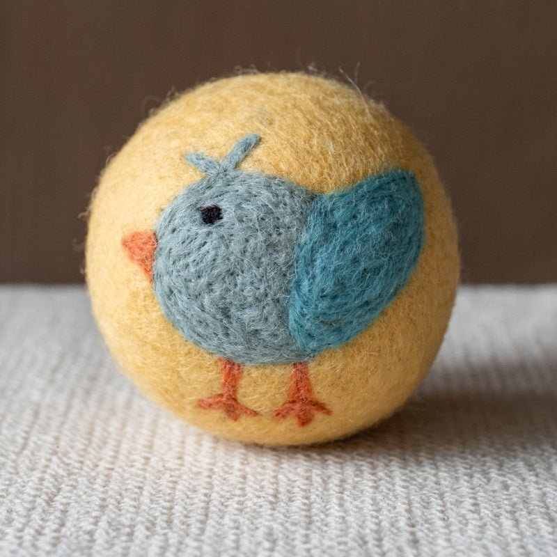 A yellow felted wool dryer ball featuring a cute bluebird design, resting on a textured beige fabric surface against a brown background.