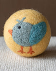 A yellow felted wool dryer ball featuring a cute bluebird design, resting on a textured beige fabric surface against a brown background.