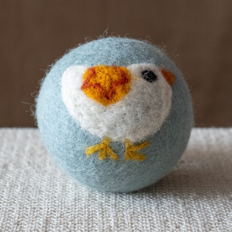 A blue felted wool dryer ball featuring a white bird with a yellow and red wing, resting on a textured beige fabric surface against a brown background.