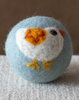 A blue felted wool dryer ball featuring a white bird with a yellow and red wing, resting on a textured beige fabric surface against a brown background.
