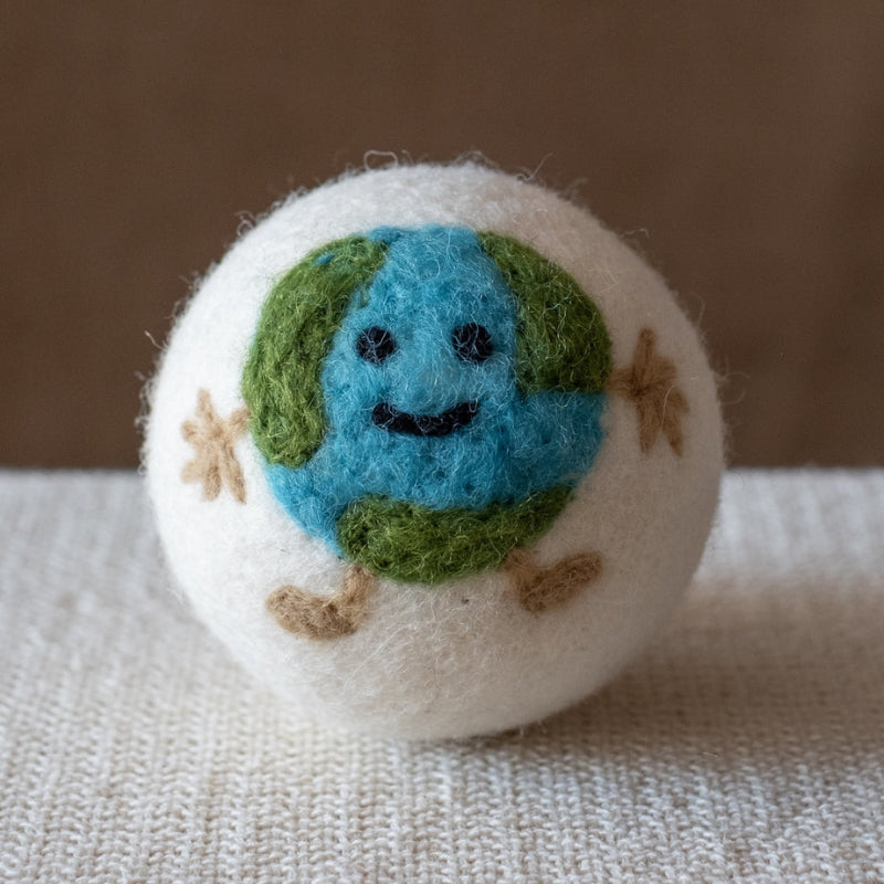 A white felted wool dryer ball featuring a Happy smiling earth design, resting on a textured beige fabric surface against a brown background.