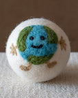 A white felted wool dryer ball featuring a Happy smiling earth design, resting on a textured beige fabric surface against a brown background.