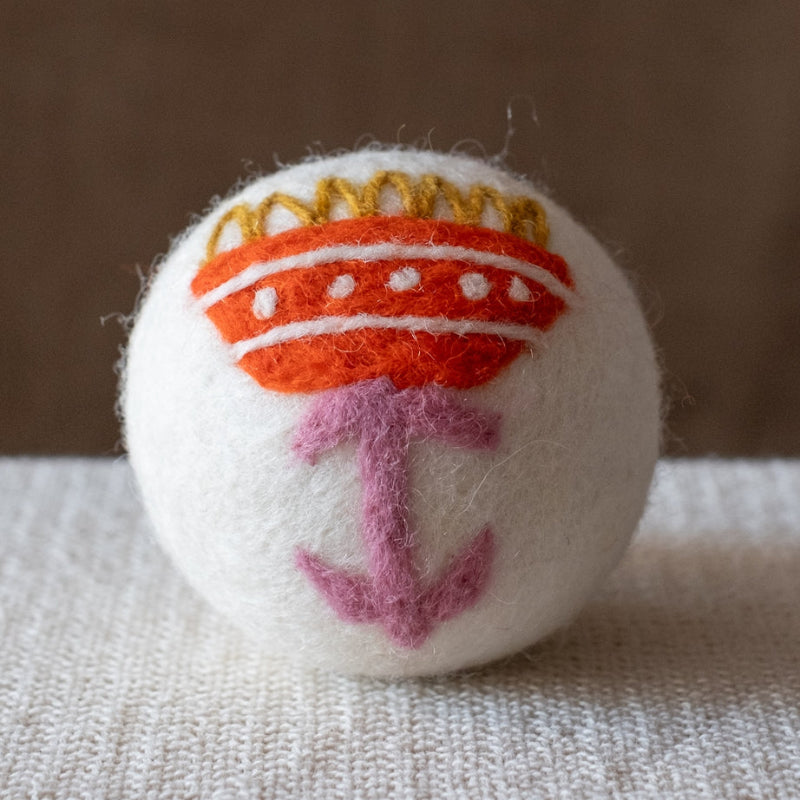 A white felted wool dryer ball featuring a orange folk art flower with a pink stem, and yellow petals, resting on a textured beige fabric surface against a brown background.