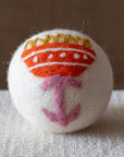 A white felted wool dryer ball featuring a orange folk art flower with a pink stem, and yellow petals, resting on a textured beige fabric surface against a brown background.