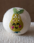 A white felted wool dryer ball featuring a happy green pear design, resting on a textured beige fabric surface against a brown background.