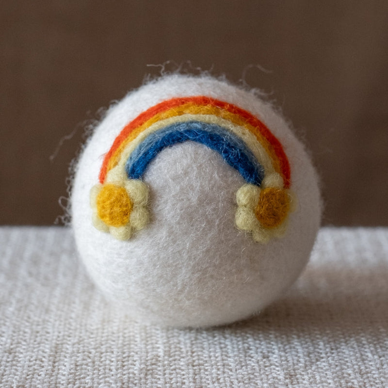 A white felted wool dryer ball featuring a rainbow ending in raised yellow daisies, resting on a textured beige fabric surface against a brown background.