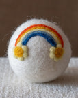 A white felted wool dryer ball featuring a rainbow ending in raised yellow daisies, resting on a textured beige fabric surface against a brown background.