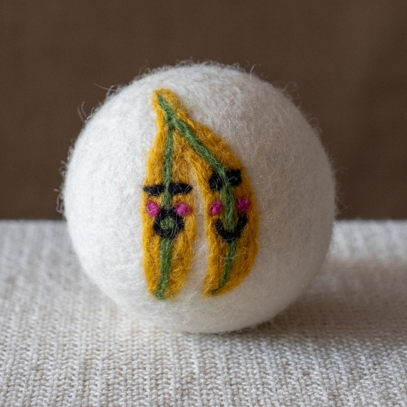 A white felted wool dryer ball with a design featuring a pair of happy bananas each with their own smiley face,  resting on a textured beige fabric surface against a brown background.