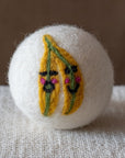 A white felted wool dryer ball with a design featuring a pair of happy bananas each with their own smiley face,  resting on a textured beige fabric surface against a brown background.