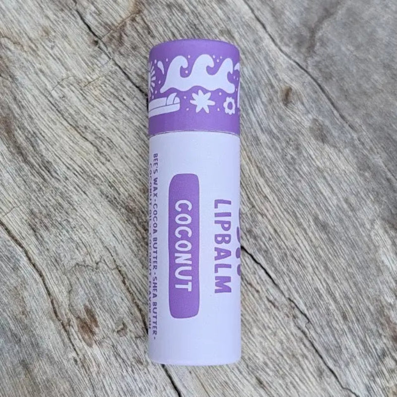 A coconut Hawaiian Lip balm in a sustainable white paper tube with purple designs on it, sitting on a wooden surface.