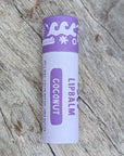 A coconut Hawaiian Lip balm in a sustainable white paper tube with purple designs on it, sitting on a wooden surface.