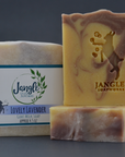 Jangle's Lovely Lavender Soap. The yellow swirled with purple bar is stamped with an image of a goat kicking bubbles. Two bars are unwrapped and one is upright on top of the other and the third bar is wrapped in paper. 