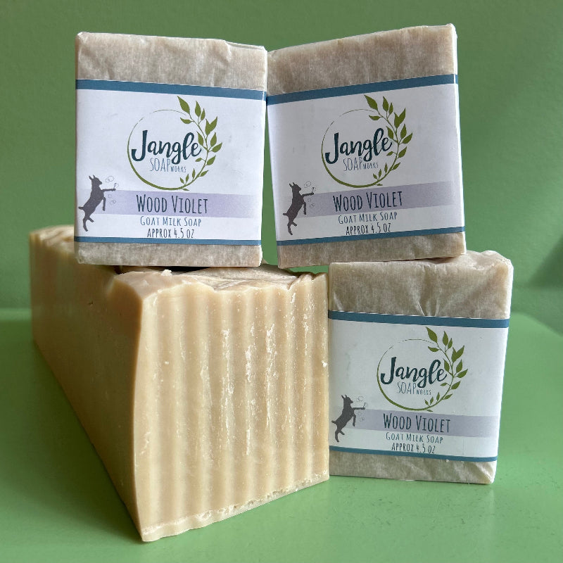 Three bars of jangle soap works wood violet soap on a green background with a bulk loaf of the creamy colored goat milk soap. 