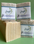 Three bars of jangle soap works wood violet soap on a green background with a bulk loaf of the creamy colored goat milk soap. 