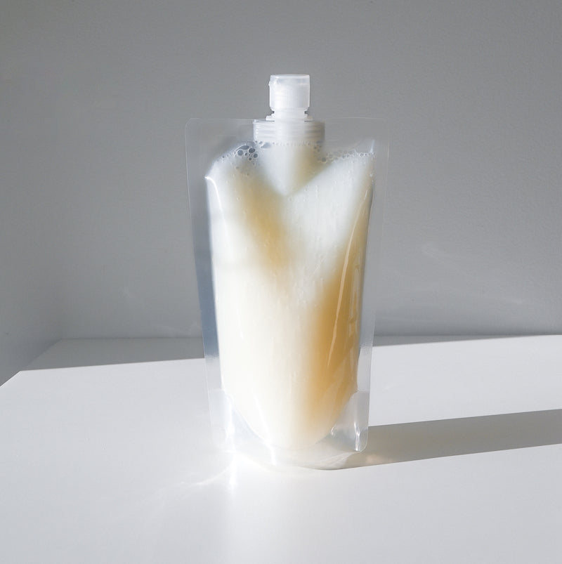 A clear plastic returnable pouch of leave in conditioner with a screw cap stands upright on a white surface, casting a shadow behind it. The pouch is filled with a creamy white liquid.