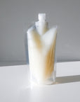 A clear plastic returnable pouch of leave in conditioner with a screw cap stands upright on a white surface, casting a shadow behind it. The pouch is filled with a creamy white liquid.
